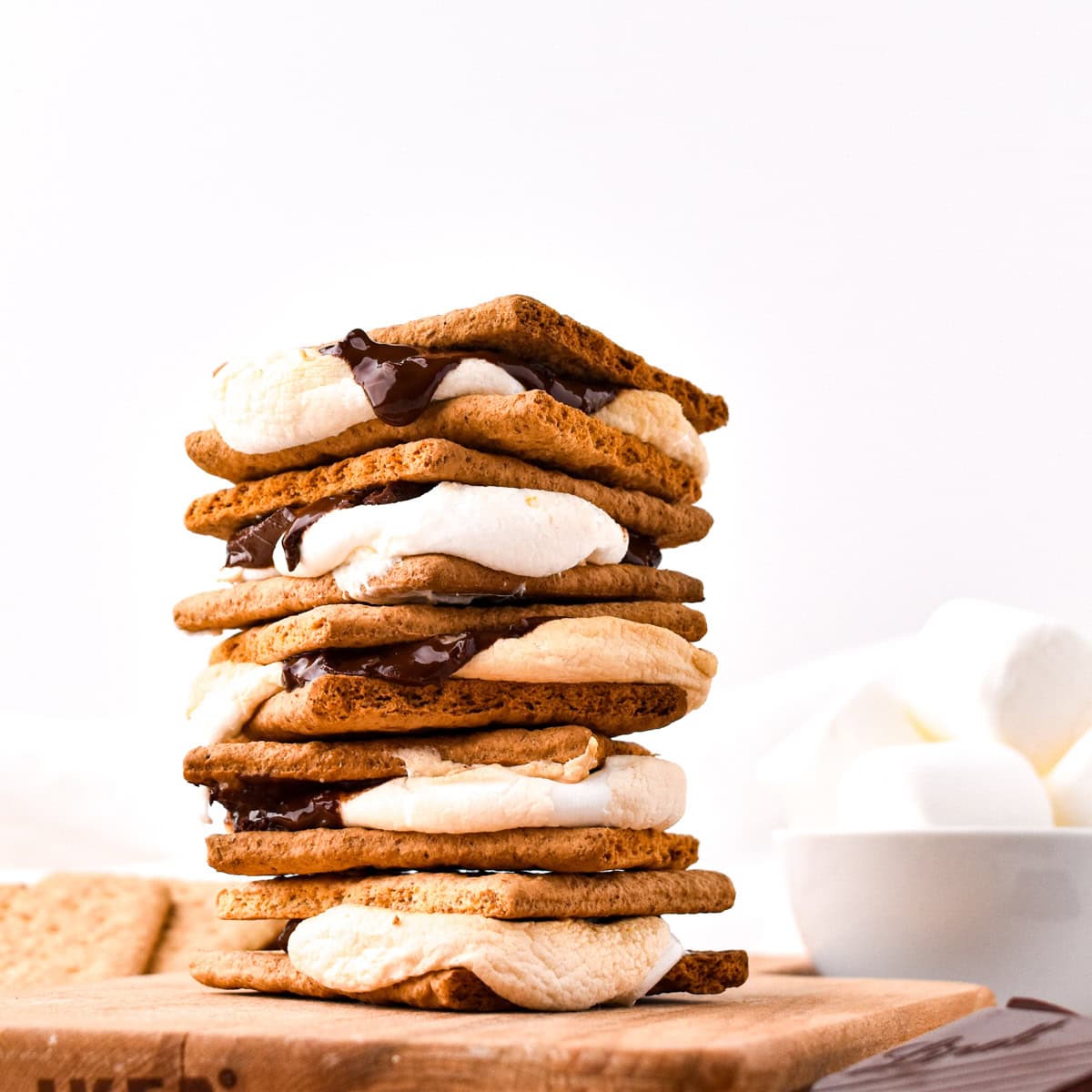 Stack of smores on wood board with bowl of marshmallows in background.