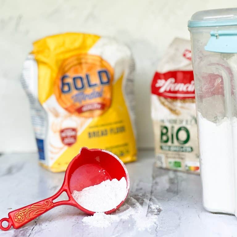 Orange measuring cup with all-purpose flour spilling onto table with bags of flour in background.