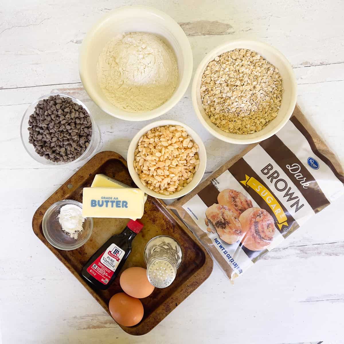 Flour, oats, rice krispies in bowls and bag of brown sugar, eggs, salt and butter on baking sheet.