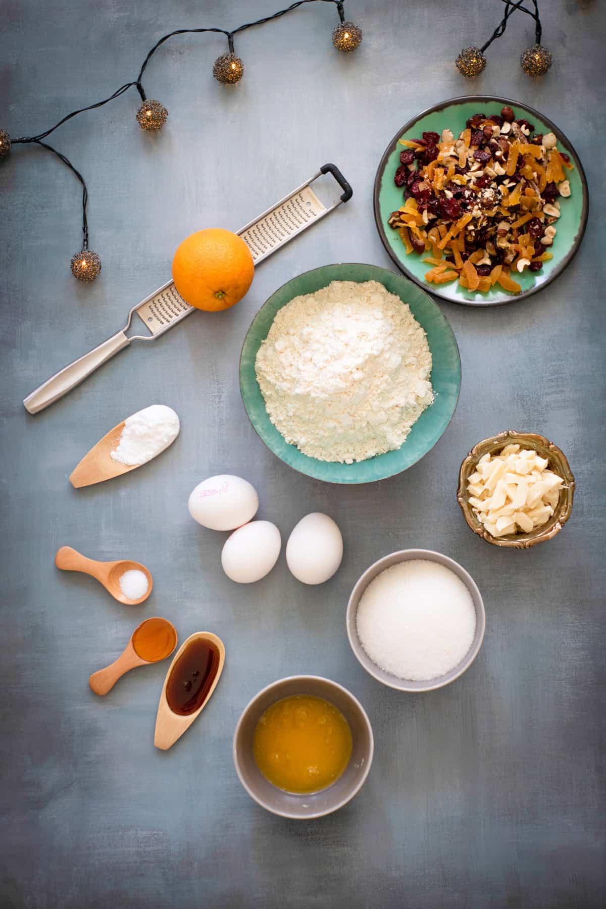 Flour, eggs, dried fruit, orange with grater, white chocolate on blue surface.